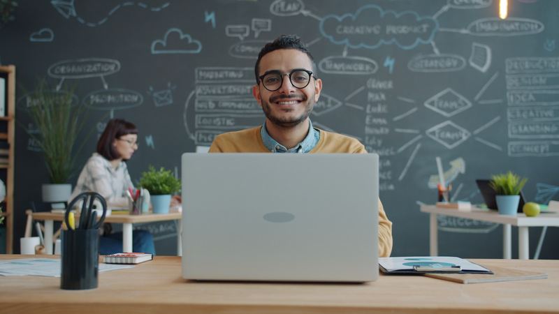 A business owner smiling at his laptop with a wall of plans behind him