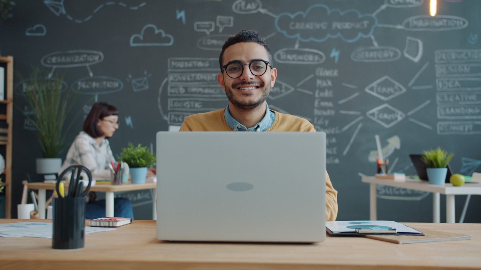 A business owner smiling at his laptop with a wall of plans behind him